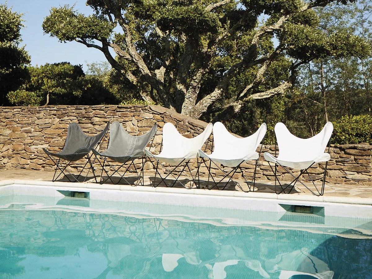 Fauteuils AA Airborne aux housses écrues et grises alignés au bord d’une piscine devant un mur en pierre