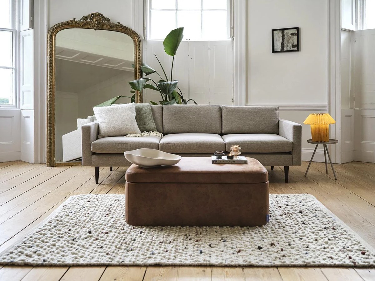Canapé en tissu clair, pouf en cuir et tapis bouclé dans un salon lumineux au parquet chêne